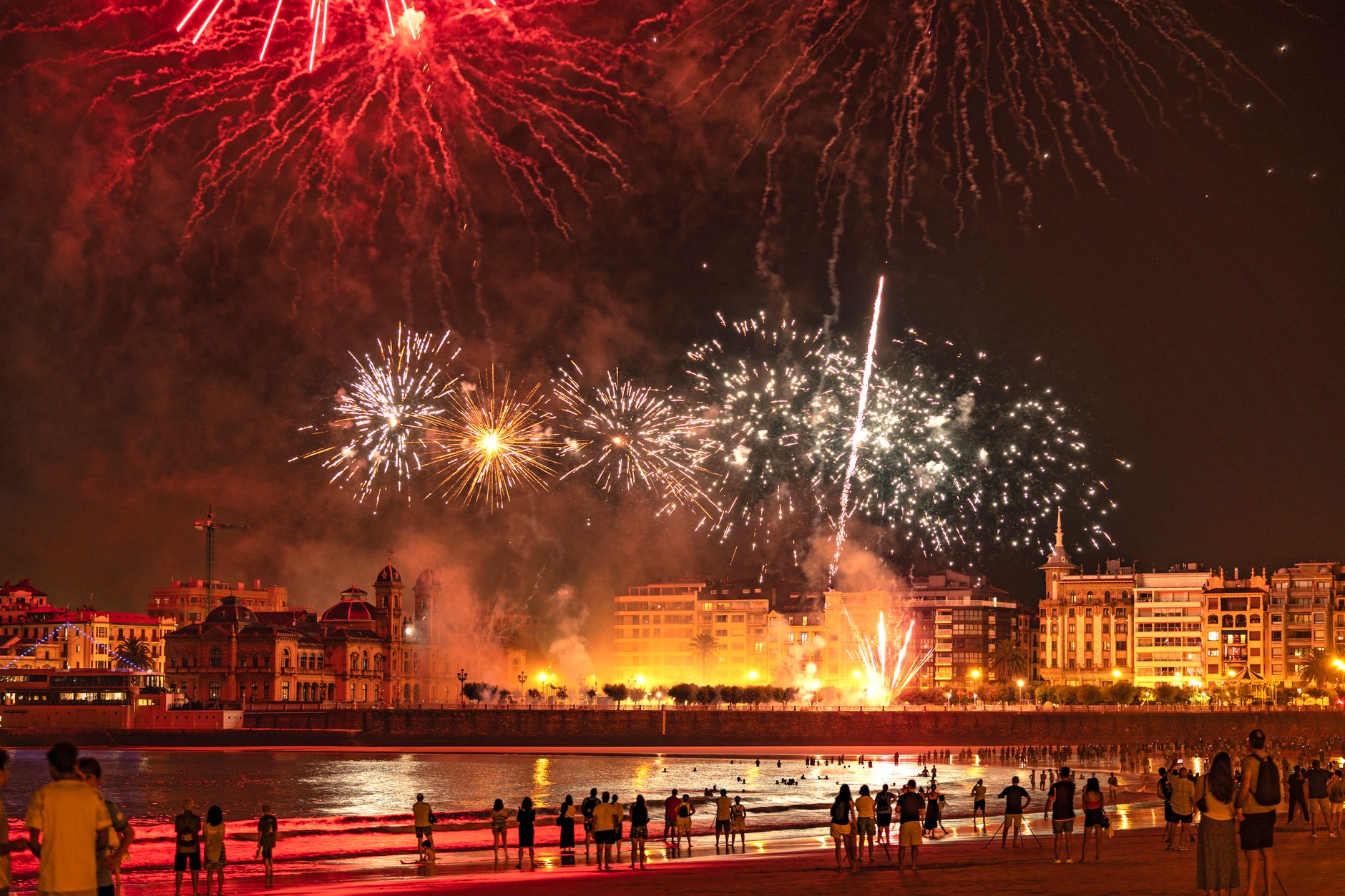 Bright fireworks illuminate a coastal cityscape at night, drawing an audience along the beach.